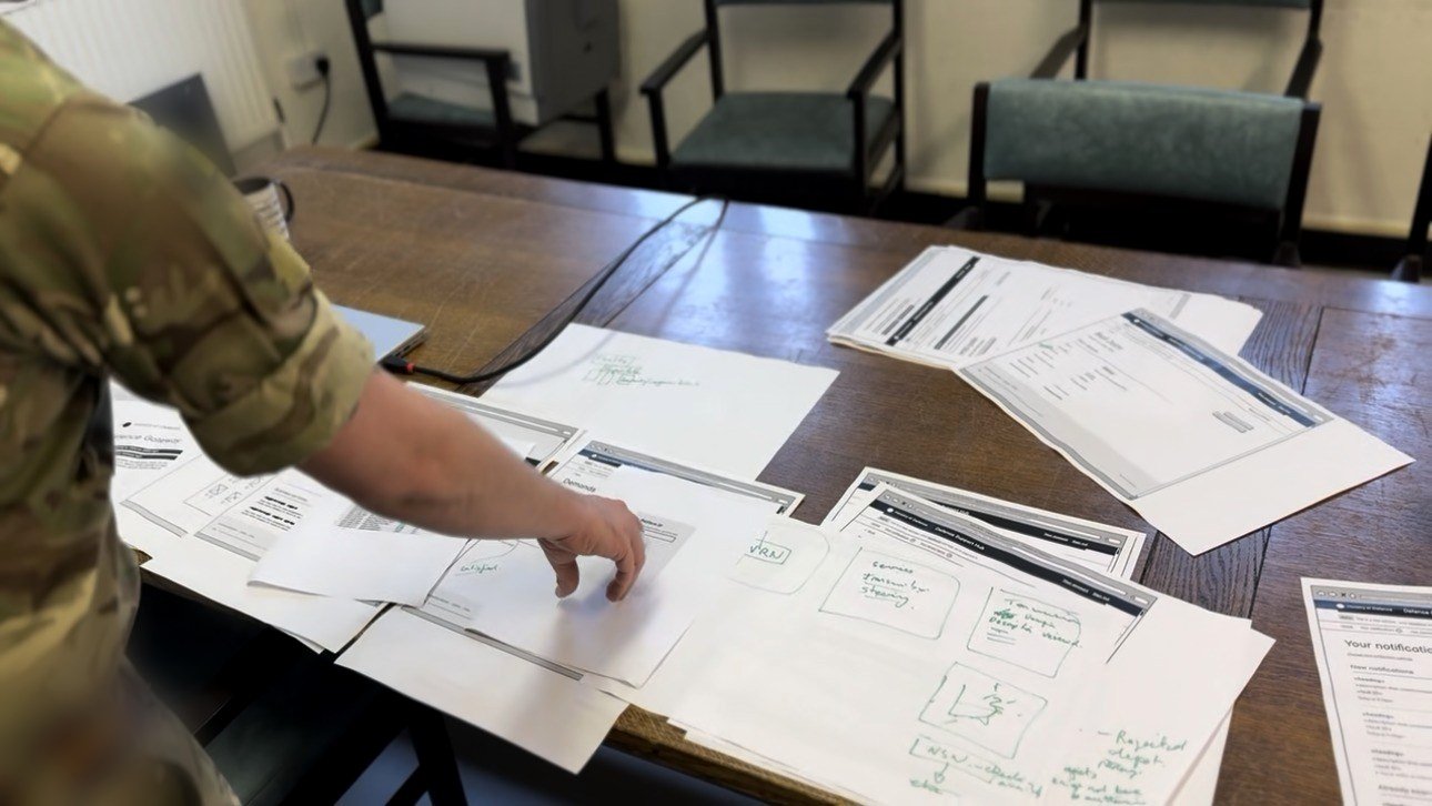 A soldier's hand moving paper prototype screens spread across a briefing-room table